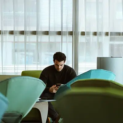 Cristina Lain Una persona está sentada sola en una habitación moderna, con una luz tenue y grandes ventanales. Observa un dispositivo, rodeado de sillones verdes y verde azulado. La luz se filtra a través de cortinas transparentes, creando una atmósfera tranquila. Branding Fotografía Mentorias