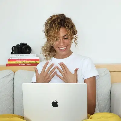 Cristina Lain Una persona con cabello rizado está sentada en un sofá, sonriendo y mirando la pantalla de un portátil. En el estante que hay detrás hay una pila de libros y una cámara. El ambiente es informal y hogareño, con una atmósfera relajada. Branding Fotografía Mentorias