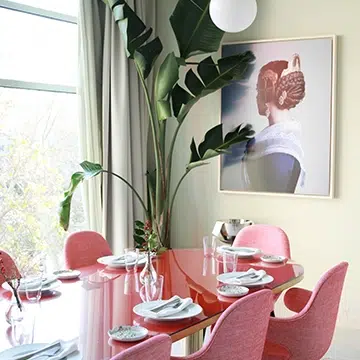 Cristina Lain Un comedor moderno con una mesa roja brillante y sillas rosas, con platos y vasos. Una gran planta y un retrato enmarcado decoran el rincón. La luz entra a raudales por una ventana con cortinas blancas transparentes. Branding Fotografía Mentorias