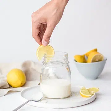 Cristina Lain Una mano sostiene una rodaja de limón sobre un tarro de leche en un plato blanco. Dos rodajas de limón descansan sobre el plato cerca de una cuchara. Un cuenco lleno de limones y un limón entero se encuentran en el fondo sobre una superficie blanca. Branding Fotografía Mentorias