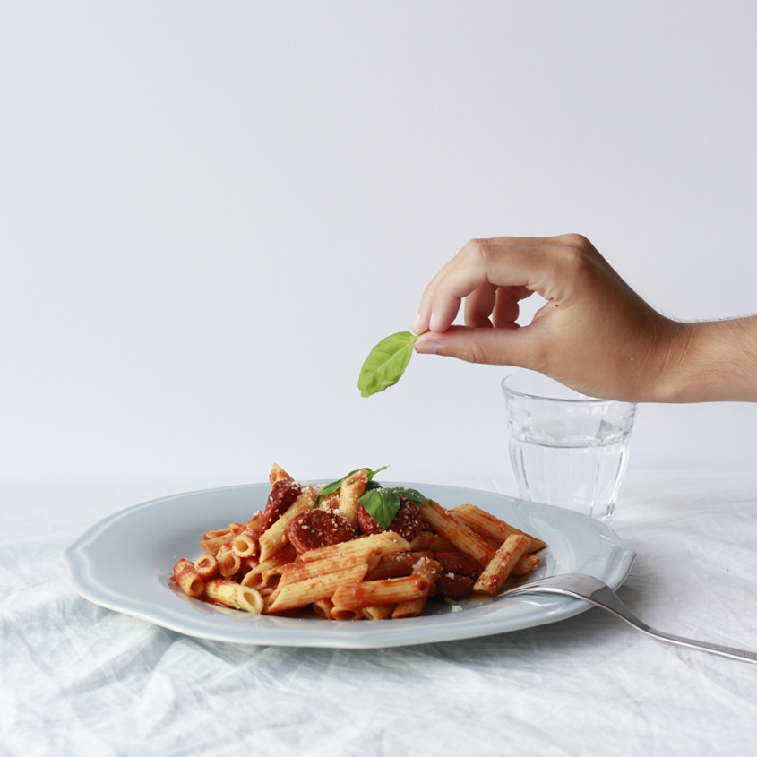 Cristina Lain Una mano sostiene una hoja de albahaca sobre un plato de pasta penne con salsa de tomate, adornado con queso rallado en un plato azul claro. Un tenedor yace sobre la mesa y un vaso de agua al lado del plato. Branding Fotografía Mentorias