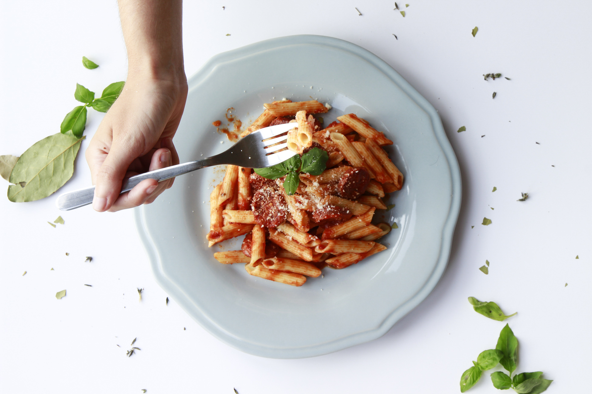 Cristina Lain Un plato de pasta penne con salsa de tomate y queso rallado, adornado con hojas de albahaca. Una mano sostiene un tenedor, lista para dar un mordisco. Albahaca fresca y trocitos de especias se encuentran esparcidos por el plato sobre una superficie blanca. Branding Fotografía Mentorias