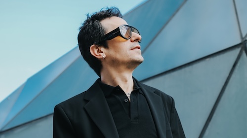 Cristina Lain Una persona con cabello corto y oscuro, con grandes gafas de sol y un abrigo negro, adopta una pose que recuerda a una fotografía sobre un fondo arquitectónico moderno bajo un cielo despejado. Branding Fotografía Mentorias