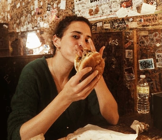 Cristina Lain Una persona sentada a la mesa de un restaurante con poca luz, con un jersey verde, posa de forma divertida con una hamburguesa al estilo Cris Laín. La pared de madera de detrás está cubierta de grafitis, lo que le da un toque urbano, mientras que sobre la mesa hay una botella de agua. Branding Fotografía Mentorias