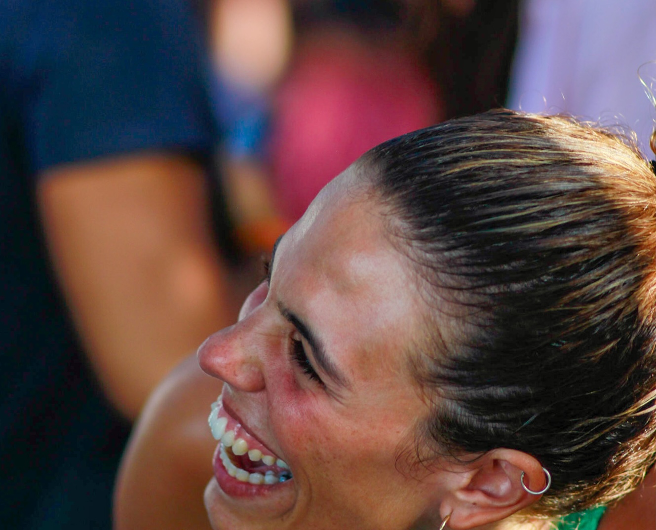 Cristina Lain Una mujer con el pelo largo recogido se ríe a carcajadas en un evento al aire libre, lo que recuerda a una escena captada por Cris Laín. Lleva pendientes y está rodeada por una multitud borrosa, lo que sugiere la atmósfera animada que suele captar. Su expresión transmite alegría y diversión. Branding Fotografía Mentorias