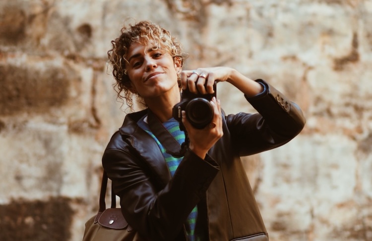 Cristina Lain Una persona de pelo rizado, que recuerda al estilo de Cris Laín, sostiene una cámara mientras sonríe y viste una chaqueta de cuero sobre una camisa de rayas. Al fondo se ve una pared de piedra con textura. Branding Fotografía Mentorias