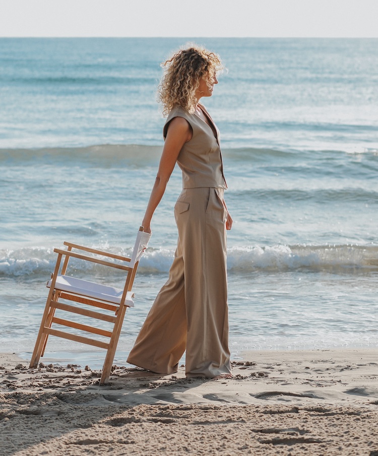 Cristina Lain Una mujer de cabello rizado se encuentra de pie en una playa de arena, con una camiseta sin mangas de color beige y pantalones anchos. Sostiene una silla plegable de madera, que recuerda el arte sereno de Cris Laín, y contempla las tranquilas olas del océano bajo un cielo despejado. Branding Fotografía Mentorias