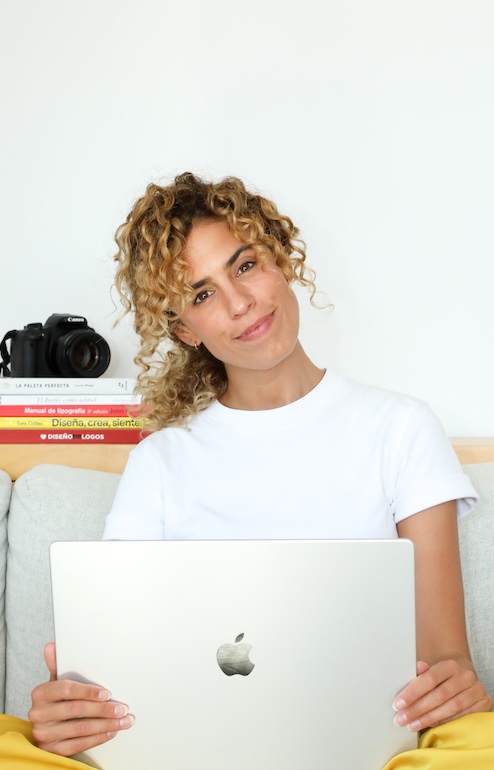 Cristina Lain Una persona de cabello rizado, con una camisa blanca, sonríe mientras está sentada en un sofá sosteniendo una computadora portátil abierta, lista para recibir mentorías personalizadas. En el fondo hay libros y una cámara apilados, lo que sugiere creatividad y aprendizaje. Branding Fotografía Mentorias