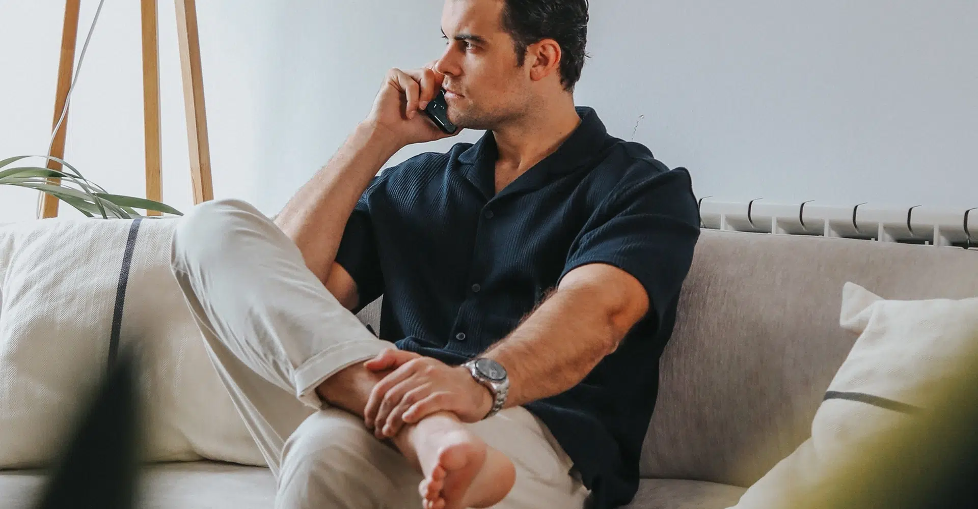 Cristina Lain Un hombre con camisa azul marino y pantalones claros está sentado descalzo en un sofá beige y habla por teléfono. Tiene las piernas cruzadas y parece relajado. Hay plantas y una lámpara de madera al fondo, contra una pared blanca. Branding Fotografía Mentorias