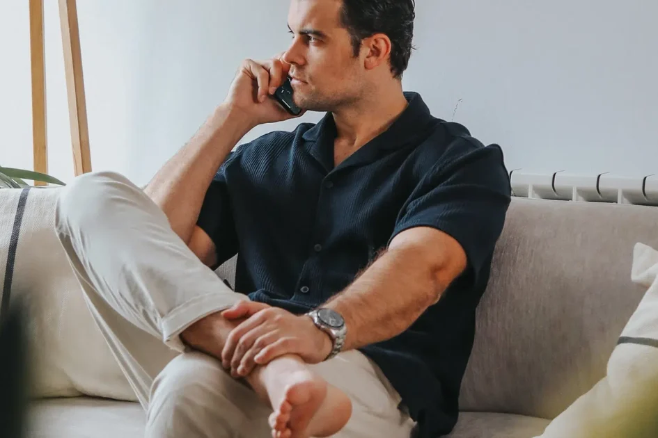 Cristina Lain Un hombre con camisa azul marino y pantalones claros está sentado descalzo en un sofá beige y habla por teléfono. Tiene las piernas cruzadas y parece relajado. Hay plantas y una lámpara de madera al fondo, contra una pared blanca. Branding Fotografía Mentorias