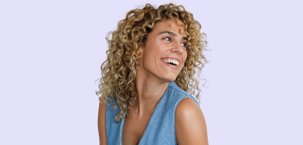 Cristina Lain Una persona con cabello rubio rizado se asemeja a la energía vibrante de una obra maestra de Cris Laín mientras sonríe y mira hacia un lado contra un fondo violeta claro, vestida con una blusa vaquera sin mangas. Branding Fotografía Mentorias